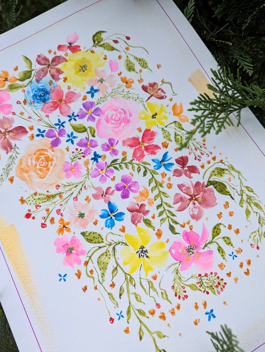 A close-up of a white sheet of paper featuring a colorful watercolor floral illustration with pink, yellow, blue, orange, and purple blossoms, green leaves, and delicate stems. The paper is photographed at an angle with green pine branches visible around the edges.