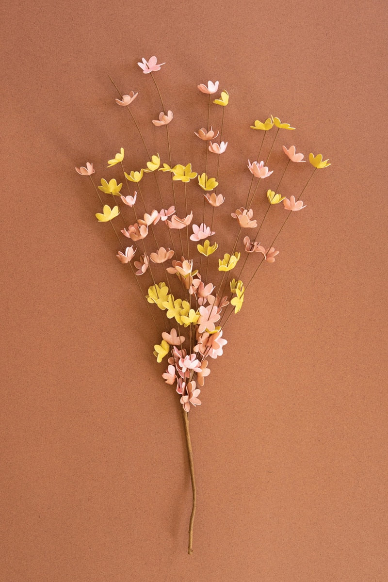 A decorative artificial flower stem made of many small paper-like blossoms in pastel pink, peach and yellow on thin twigs, set against a solid warm brown background