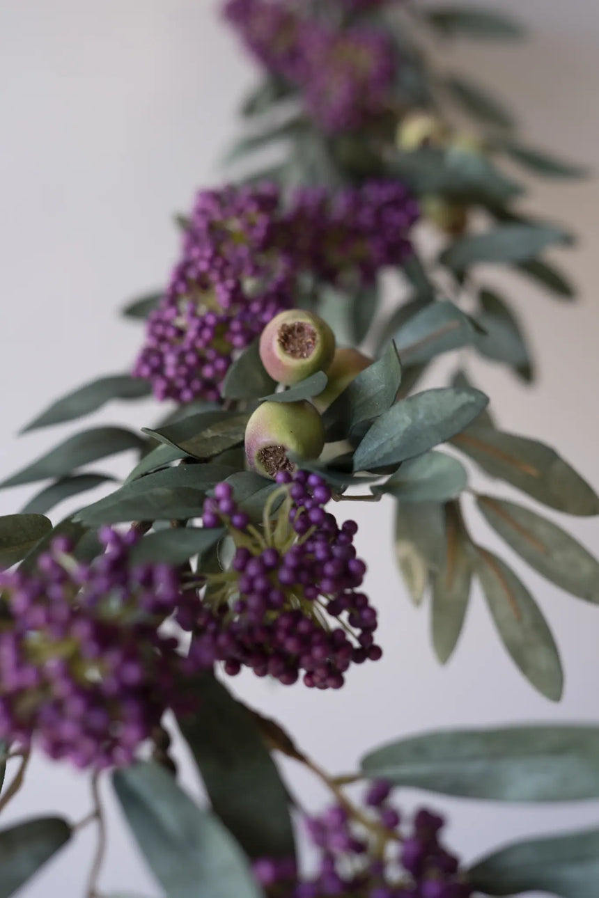 Artificial floral garland with clusters of small purple berries, muted green leaves, and two faux green berries, shown against a plain light background.