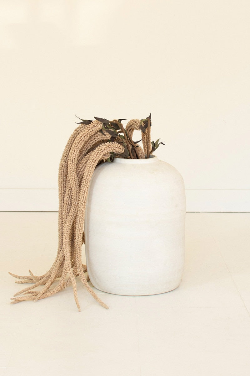 A matte white ceramic vase with dried decorative branches and long beige braided hair draped over the side, set against a plain off-white background.