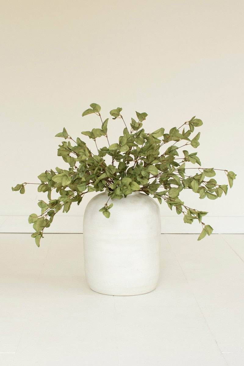 A white ceramic vase filled with green trailing eucalyptus branches, set against a clean, off-white background.