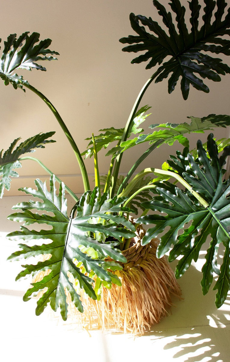 A potted artificial Philodendron plant with large, deeply lobed green leaves, planted in a natural tan coir basket pot, set against a plain beige wall with soft sunlight and leaf shadows.