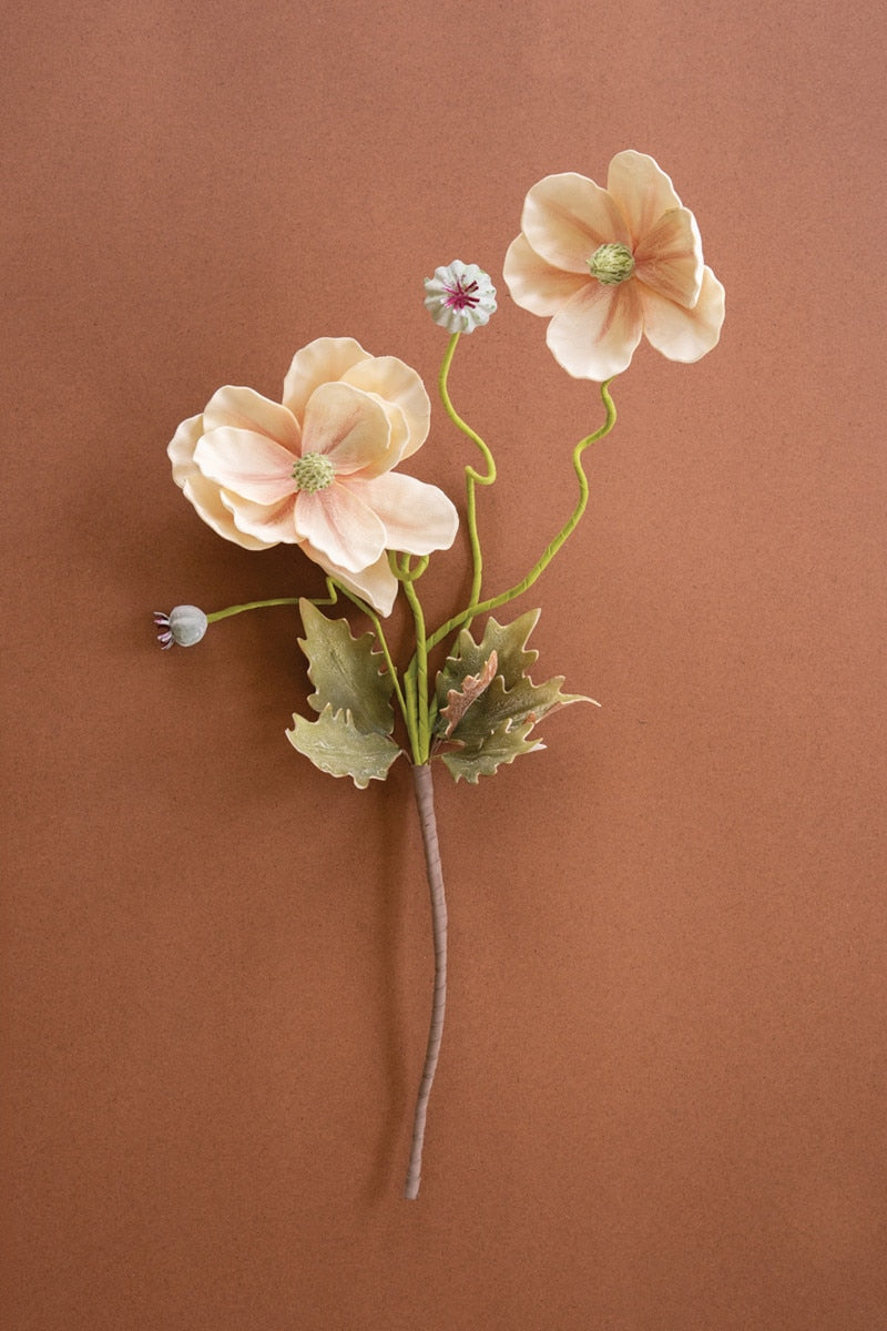 A single artificial flower stem with three pale peach blossoms, green stems and leaves, and small buds, arranged on a warm brown background.