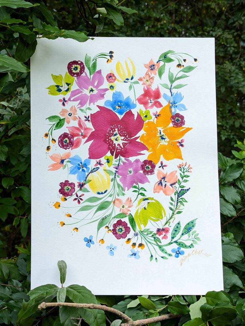 An original watercolor painting featuring a vibrant watercolor floral design with pink, blue, yellow, orange and purple blossoms, displayed outdoors against a background of green leaves and foliage.