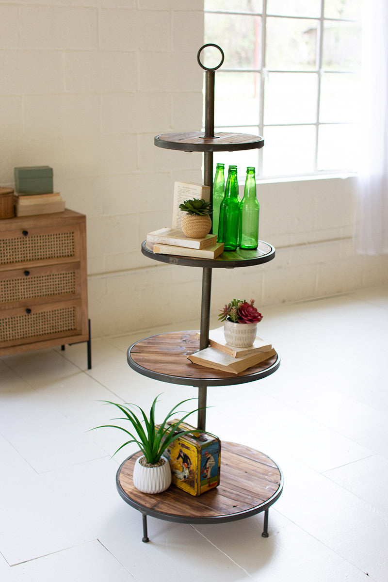 A four-tiered display tower with recycled wood and metal shelves, standing in a bright room with various items placed on the shelves.