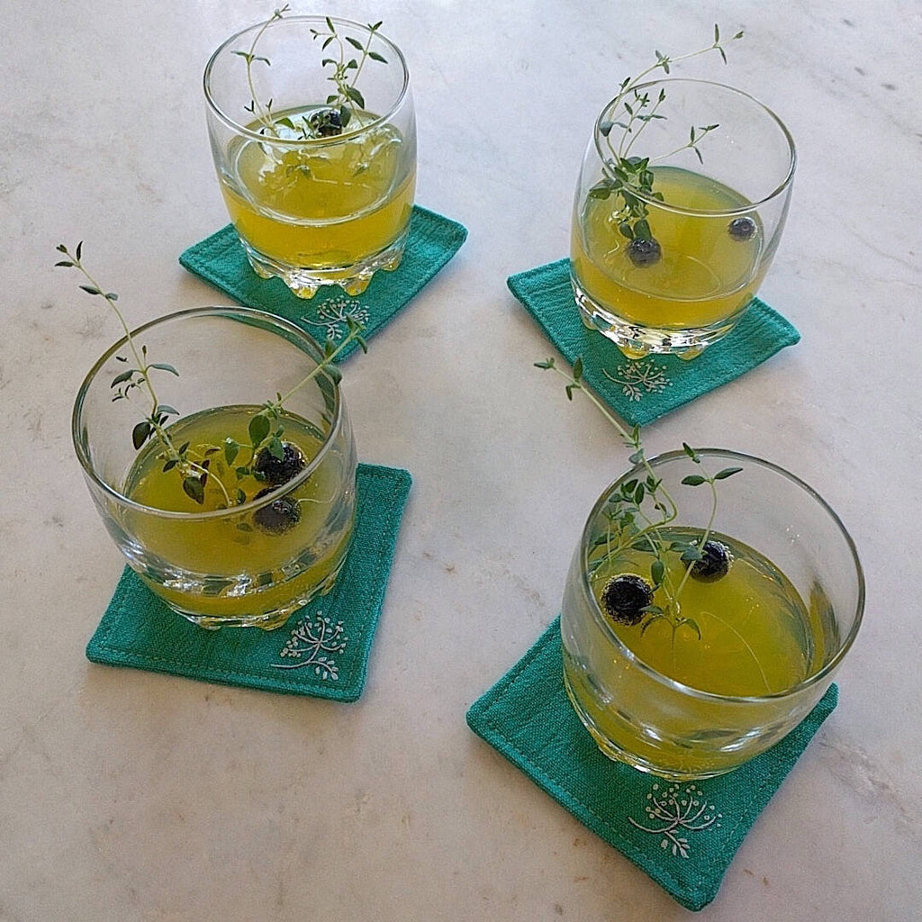 4 glasses filled with mojito with thyme leaves and blueberries atop moss green linen coasters