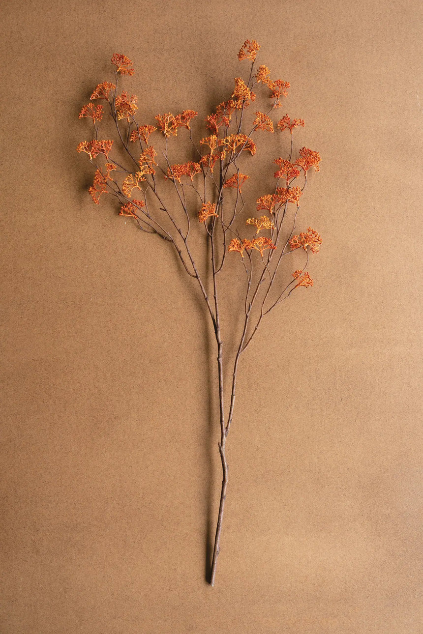 A single dried floral stem with small orange flowers and thin brown branches, arranged upright against a plain warm tan background.