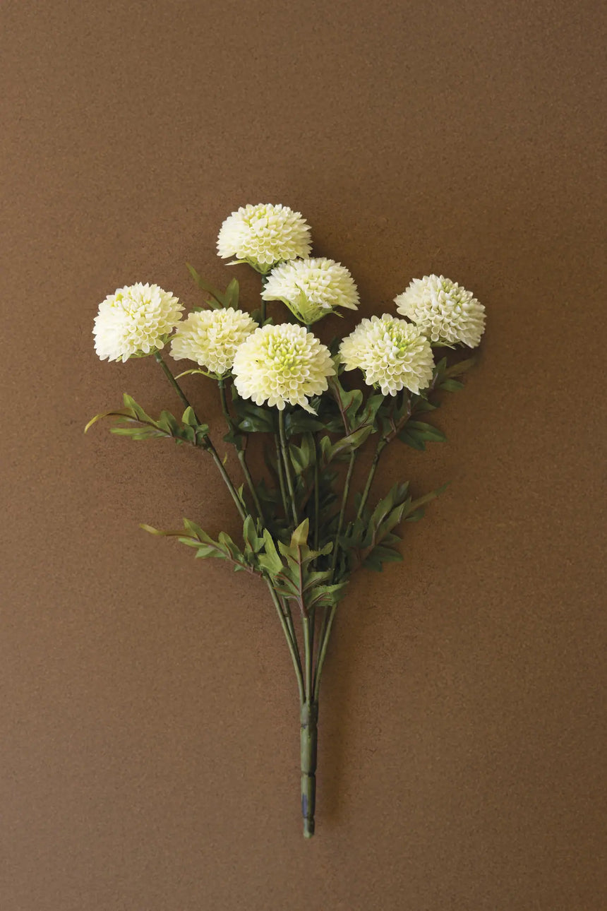 A bouquet of artificial white dalhia flowers with green stems and leaves, arranged against a solid brown background