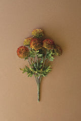 An artificial floral stem bouquet with round, textured pom-pom flowers in orange, red, and yellow tones, accented by green leaves and stems, set against a solid warm brown background
