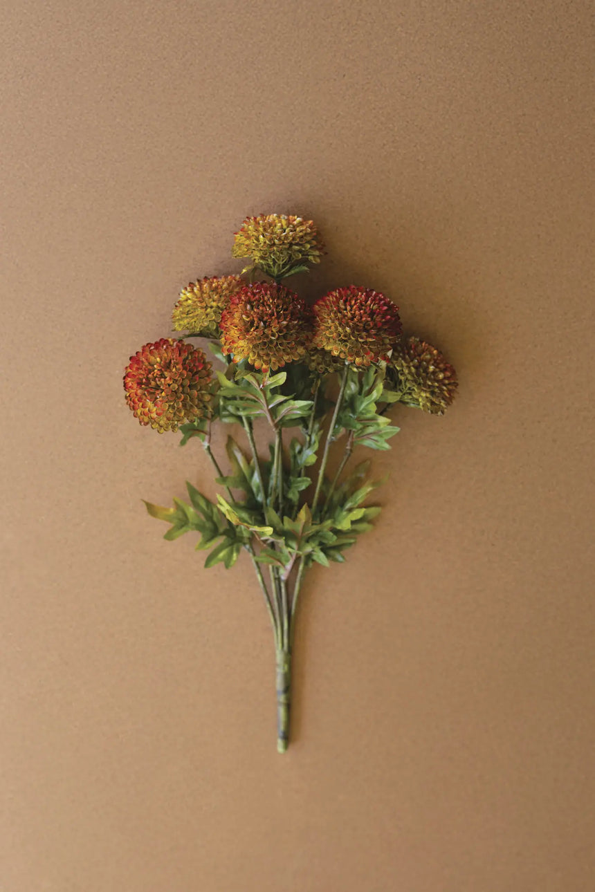 An artificial floral stem bouquet with round, textured pom-pom flowers in orange, red, and yellow tones, accented by green leaves and stems, set against a solid warm brown background