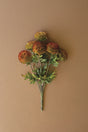 An artificial floral stem bouquet with round, textured pom-pom flowers in orange, red, and yellow tones, accented by green leaves and stems, set against a solid warm brown background