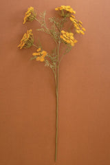 An artificial flower stem featuring clusters of small yellow-orange blossoms and delicate green-gray foliage on a long brown stem, set against a solid terracotta background.