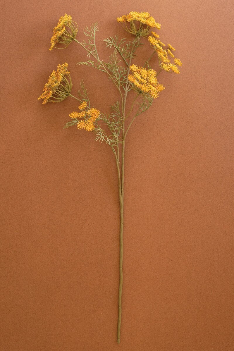 An artificial flower stem featuring clusters of small yellow-orange blossoms and delicate green-gray foliage on a long brown stem, set against a solid terracotta background.