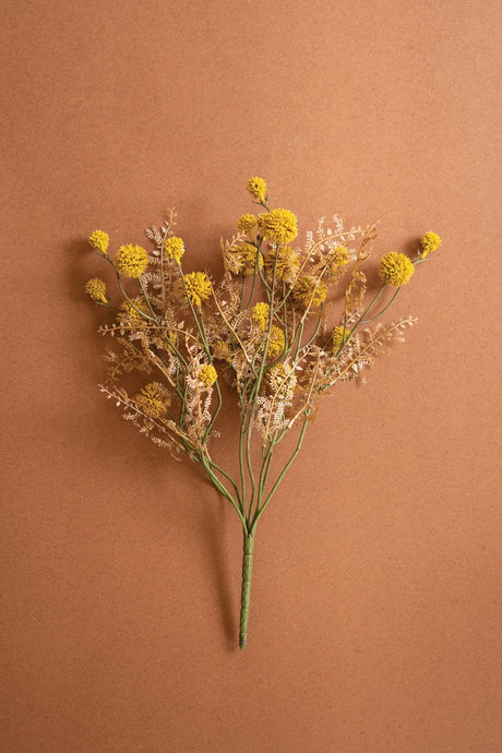 A faux dried flower bouquet with small yellow globes and pale beige filler stems on a plain warm brown background