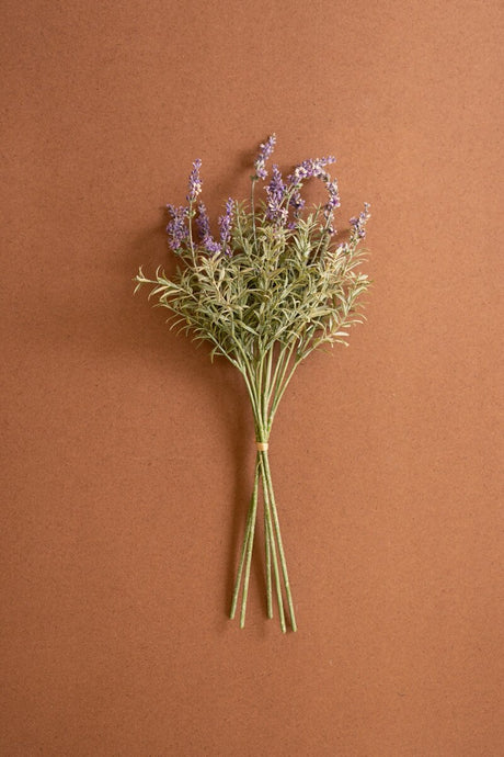 A small bouquet of artificial dried lavender stems with light green foliage and purple flowers, tied with a simple band and set against a solid warm brown background.