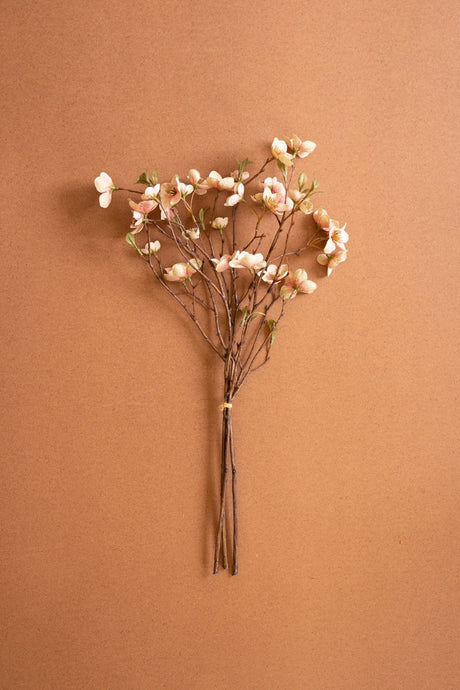A small bouquet of artificial peach blossom branches with pale pink flowers and brown stems, tied with a thin band and displayed against a solid warm brown background.