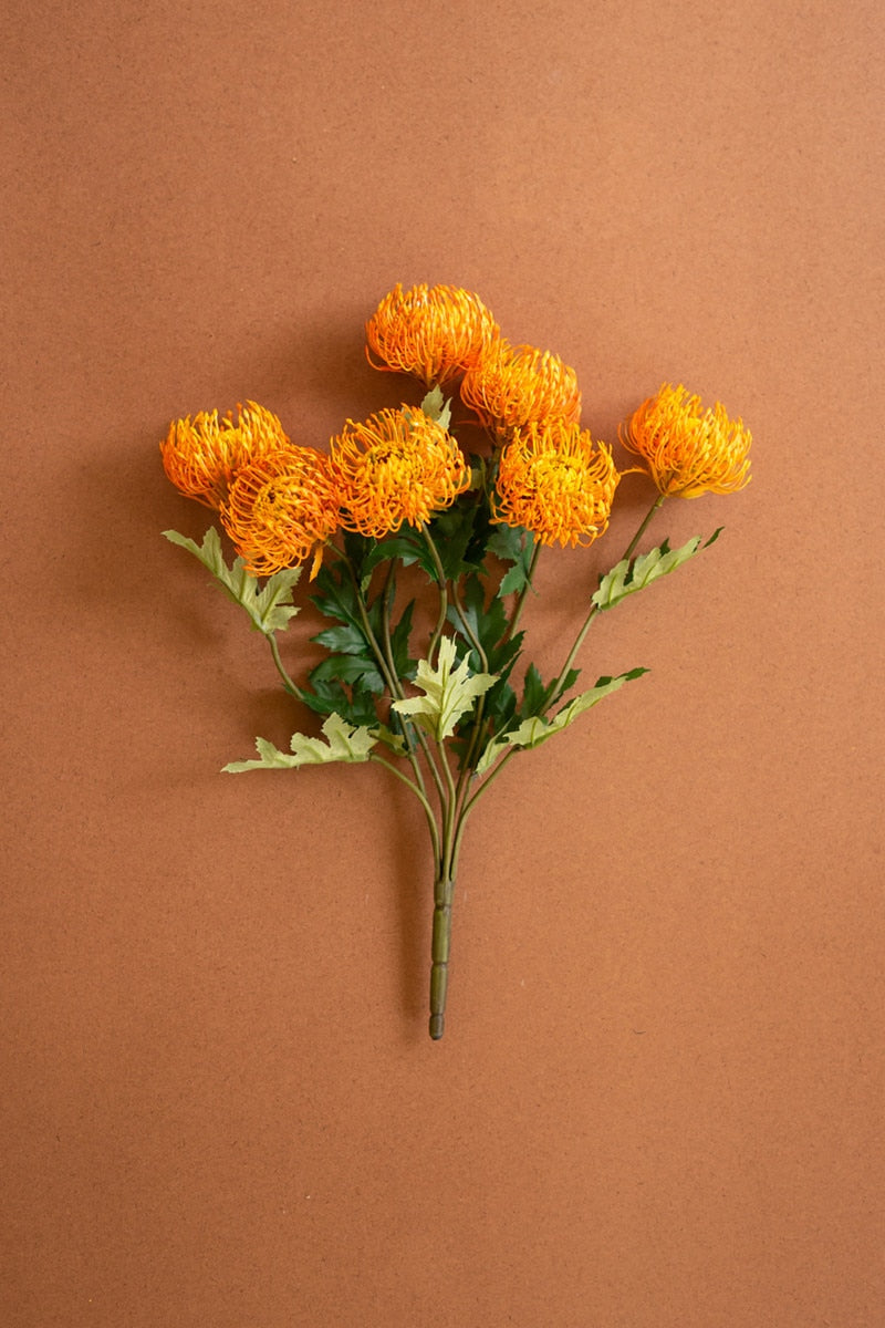 A bouquet of artificial orange protea with green leaves and stems, arranged on a solid brown background.