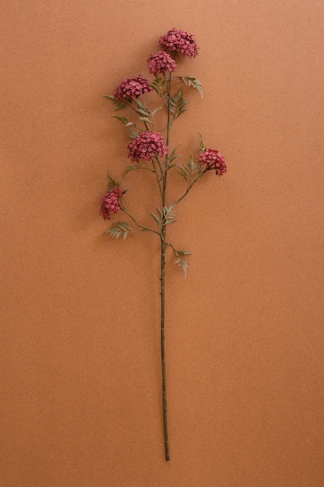 A single artificial flower stem with clusters of dusty pink blossoms and muted green leaves, displayed vertically against a solid terracotta-colored background.