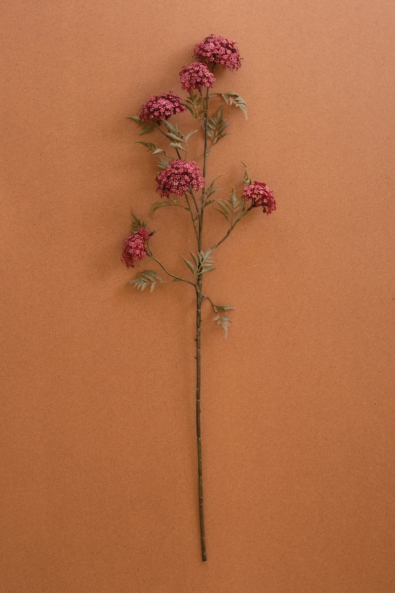 A single artificial flower stem with clusters of dusty pink blossoms and muted green leaves, displayed vertically against a solid terracotta-colored background.