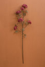 A single artificial flower stem with clusters of dusty pink blossoms and muted green leaves, displayed vertically against a solid terracotta-colored background.