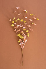 A decorative artificial flower stem made of many small paper-like blossoms in pastel pink, peach and yellow on thin twigs, set against a solid warm brown background