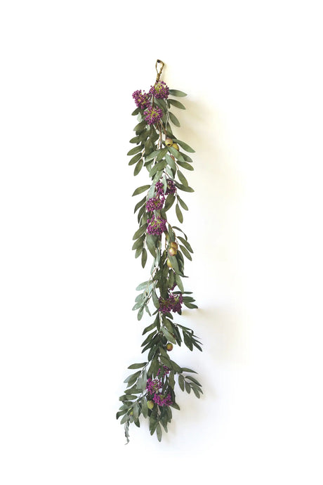 A hanging garland of olive green leaves with clusters of small purple berries and tiny gold olives, suspended against a plain white wall.