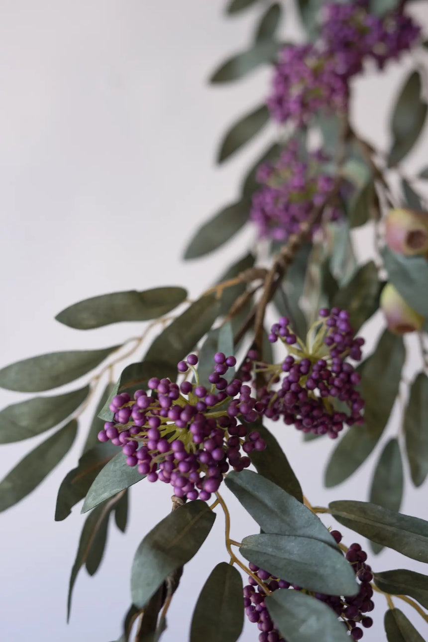 A close-up of a decorative garland featuring clusters of small purple berries and elongated olive-green leaves against a plain light background.