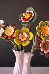 A close-up of a decorative floral arrangement made of layered eva foam flowers in yellow, orange, pink, red, white and green, placed in a pale pink ribbed vase against a dark gray background.