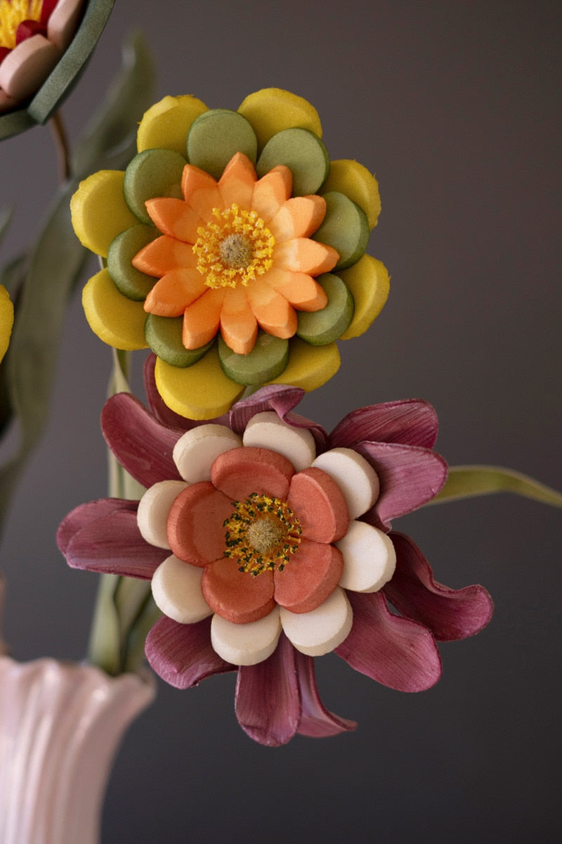 Two decorative sculptural flowers on stems against a dark gray background. The upper flower has yellow outer petals, olive green scalloped petals, and an orange center with yellow stamens.