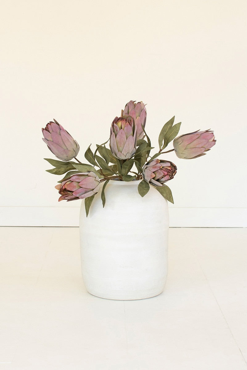 A white ceramic vase holding a bouquet of dried protea-style flowers with muted pink petals and green leaves, set against a plain off-white background.