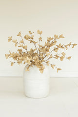 A vase arrangement featuring dried beige faux foliage with small leafy branches, displayed in a matte white ceramic vase against a clean, off-white background