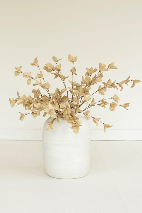 A vase arrangement featuring dried beige faux foliage with small leafy branches, displayed in a matte white ceramic vase against a clean, off-white background