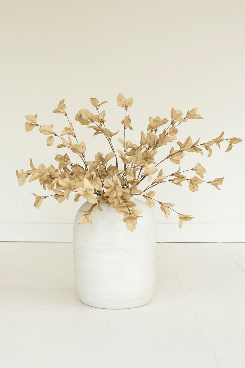 A vase arrangement featuring dried beige faux foliage with small leafy branches, displayed in a matte white ceramic vase against a clean, off-white background