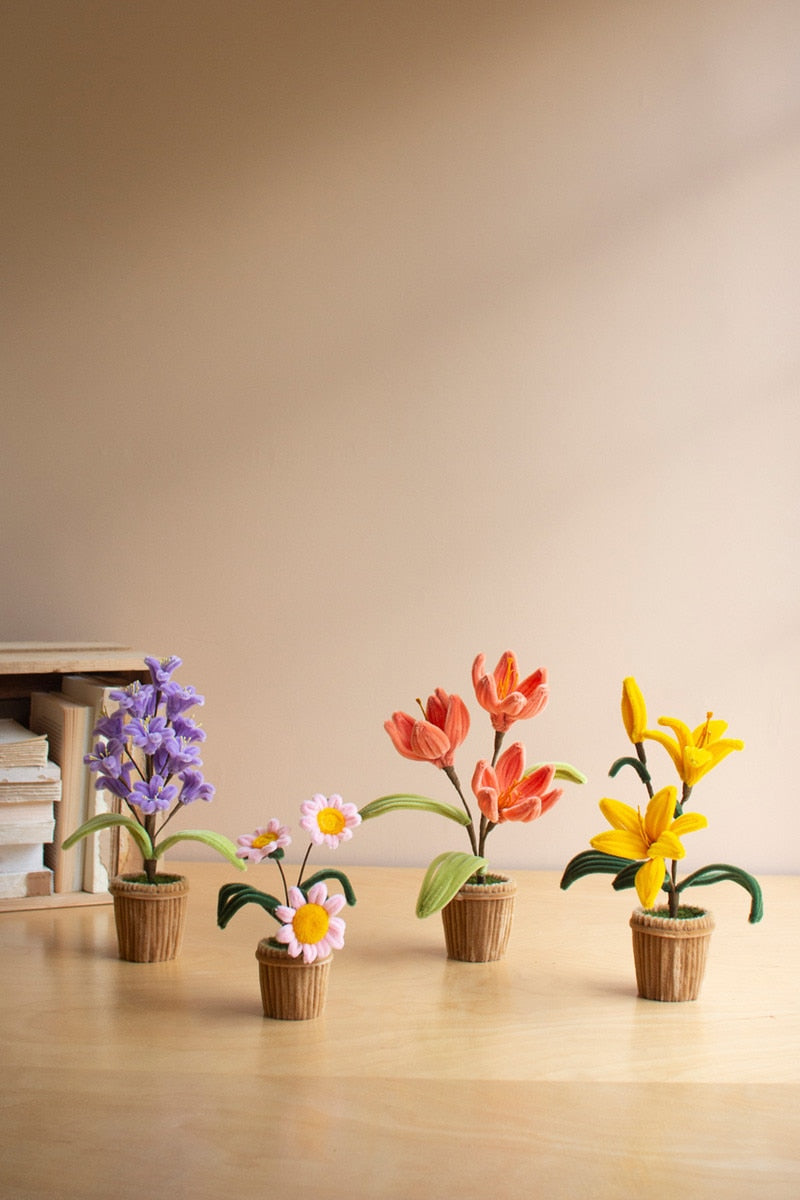 Four small artificial flower pots arranged on a light wood surface against a warm beige wall. The pots include purple hyacinths, white daisies with yellow centers, coral tulips, and yellow lilies, each in a brown woven pot.