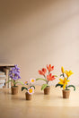 Four small artificial flower pots arranged on a light wood surface against a warm beige wall. The pots include purple hyacinths, white daisies with yellow centers, coral tulips, and yellow lilies, each in a brown woven pot.
