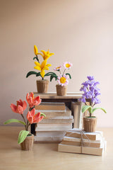 A styled arrangement of artificial flowers in woven pots, displayed on a stack of books and wrapped gift boxes against a plain beige background. The flowers include yellow lilies, pink and white daisies, purple hyacinths, and orange tulips.