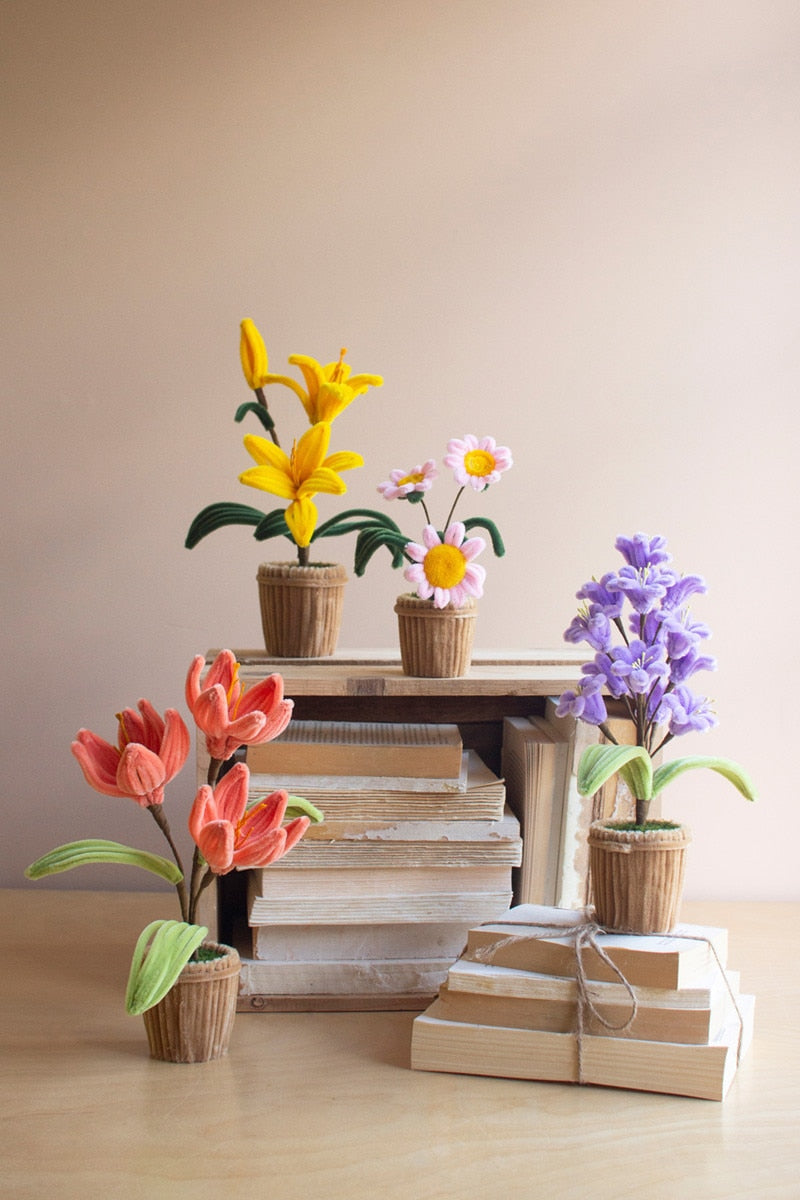 A styled arrangement of artificial flowers in woven pots, displayed on a stack of books and wrapped gift boxes against a plain beige background. The flowers include yellow lilies, pink and white daisies, purple hyacinths, and orange tulips.