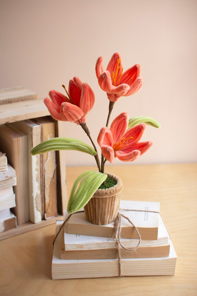 A decorative arrangement of three coral-pink lily flowers with green leaves in a small woven pot, placed on stacked books tied with twine on a wooden table.