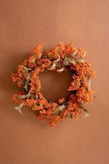 An autumn-themed floral wreath with dense clusters of orange berries and muted sage green leaves, displayed against a solid warm brown background.