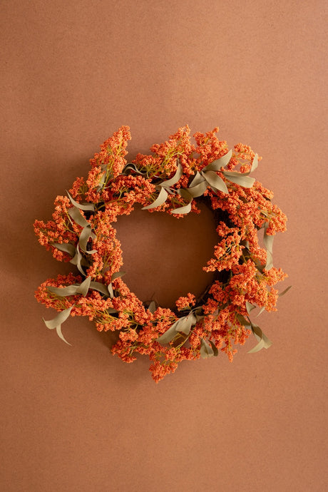 An autumn-themed floral wreath with dense clusters of orange berries and muted sage green leaves, displayed against a solid warm brown background.