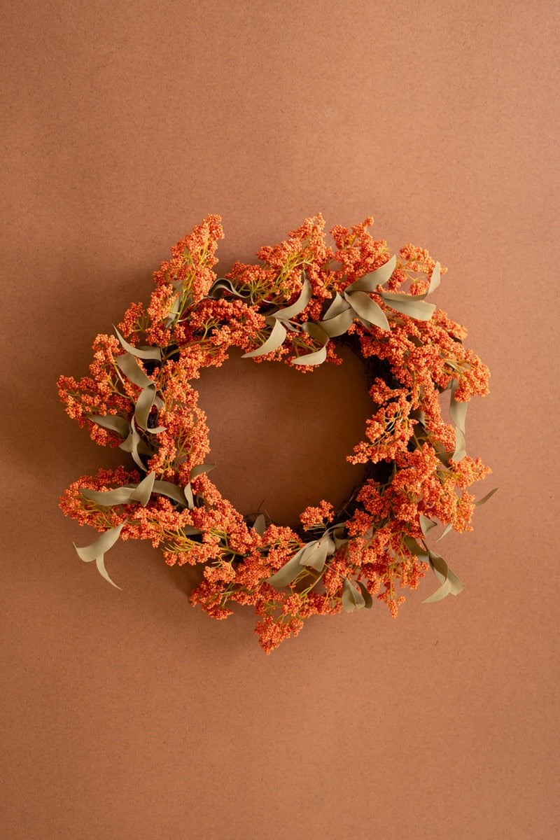 An autumn-themed floral wreath with dense clusters of orange berries and muted sage green leaves, displayed against a solid warm brown background.