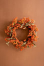 An autumn-themed floral wreath with dense clusters of orange berries and muted sage green leaves, displayed against a solid warm brown background.