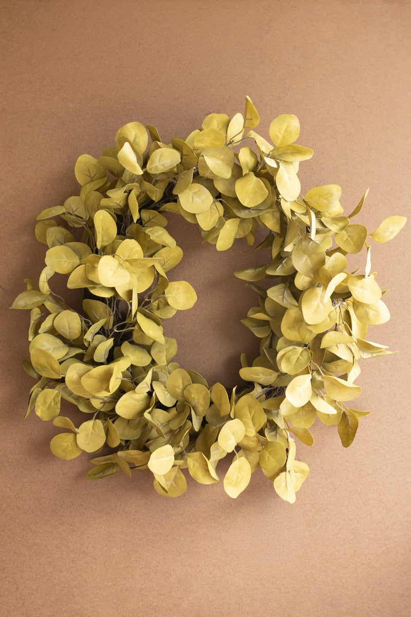 An artificial eucalyptus wreath in muted olive green and yellow-green tones, displayed against a solid brown background.