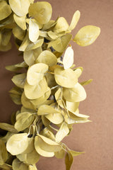 A close-up of a decorative artificial leaf garland in a muted olive-yellow hue, shown against a solid warm brown background.