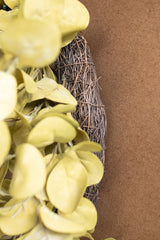 A close-up of a decorative wreath made of brown woven branches with pale yellow-green faux leaves, set against a solid brown background.