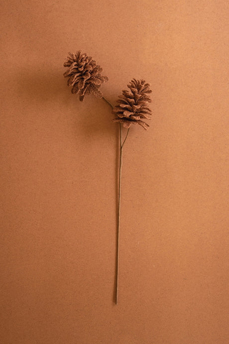 A single artificial pinecone stem decoration with two brown pinecones on a long thin stem, set against a solid warm brown background.