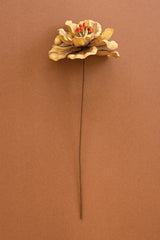 A single dried decorative artificial flower with layered tan and brown petals and a small orange-red center, attached to a long brown stem, photographed against a solid warm brown background.
