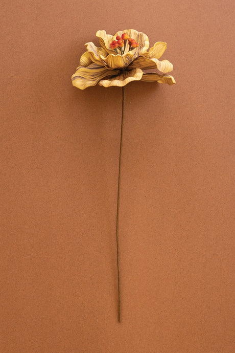A single dried decorative artificial flower with layered tan and brown petals and a small orange-red center, attached to a long brown stem, photographed against a solid warm brown background.