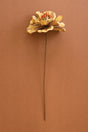 A single dried decorative artificial flower with layered tan and brown petals and a small orange-red center, attached to a long brown stem, photographed against a solid warm brown background.