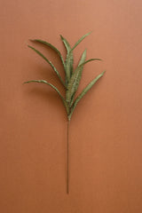 A single artificial fern stem with green fronds, laid flat on a solid terracotta brown background.